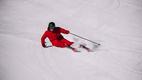 A skier on performance skis carves sharply on a groomed slope, kicking up snow.